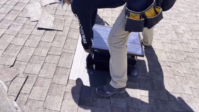 A worker installs a solar attic fan on a shingled roof, using tools and wearing a utility belt.