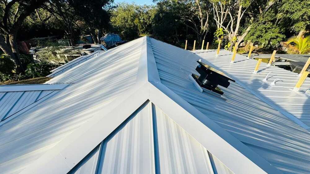 A close-up view of a metal rooftop with a central ridge, surrounded by trees and construction materials, featuring a black roof vent and vertical wooden supports.