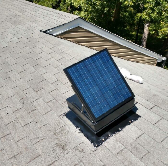 A small solar panel is installed on a sloped gray shingle roof, positioned to face the sun. The base appears sealed with black flashing material.