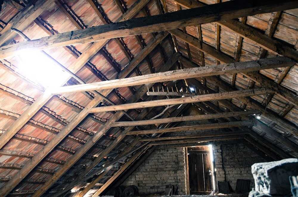 An unfinished attic with exposed wooden beams and rafters under a tiled roof, with sunlight streaming through gaps.